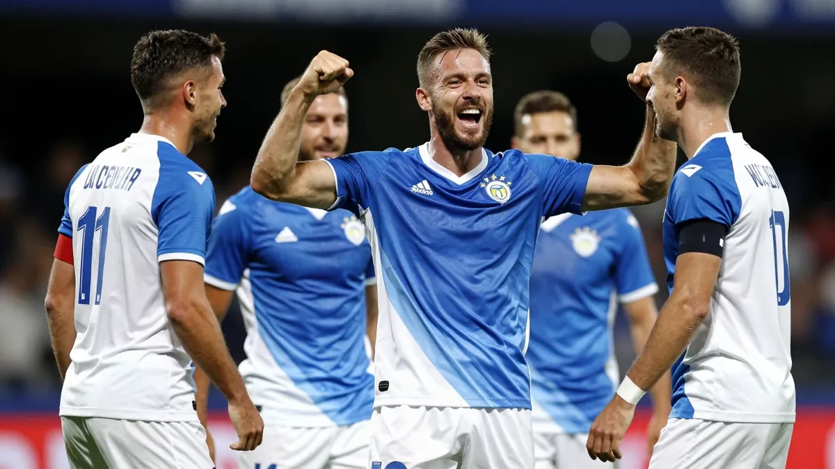 A 30-year-old Rodez player, with a muscular build, wearing a blue and white jersey, celebrating a goal with his...