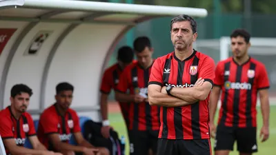 A frustrated middle-aged male coach in a red and black striped shirt, standing in a technical area, arms crossed...