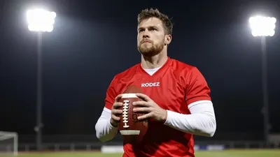 A 35-year-old Rodez player in a red jersey with white sleeves, holding a football and looking up at the floodlights with...