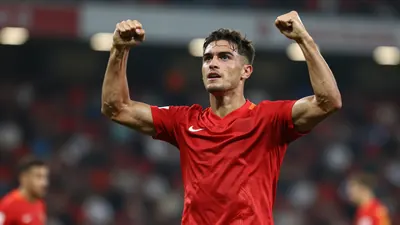 Tight action crop of a muscular 25-year-old forward in solid red kit, mid-celebration after scoring, arms raised, face...